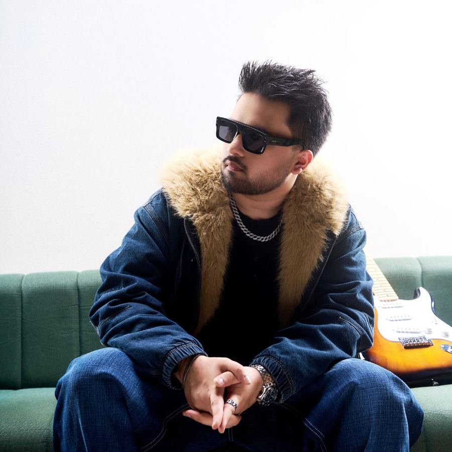 Abraham Khanna in sunglasses and a shearling-collar jacket, seated with a guitar